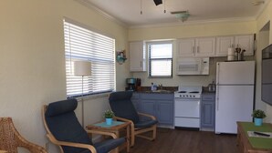 A compact kitchen with white cabinets, a stove, and a refrigerator. There are two wicker chairs and a small table with a plant. - Adorable One Bedroom One Bathroom With Easy Access To The Beach!  (Redington Beach)