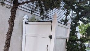 Un petit abri en bois peint en blanc, doté d’un loquet métallique, situé à côté d’un arbre et d’une clôture blanche.