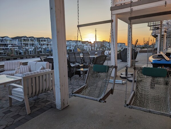 Un patio frente al mar con sillas de mimbre y una hamaca, con vistas a una marina con barcos y casas.