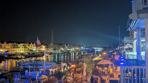 Une terrasse au bord de l’eau avec des sièges extérieurs, une vue sur le port de plaisance et un bâtiment illuminé de lumières bleues.