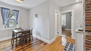 Un coin repas avec une table et des chaises en bois, une fenêtre avec des rideaux bleus et un espace cuisine avec un mur en briques et un évier.