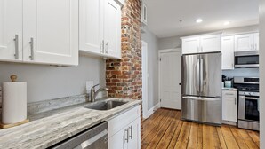 Une cuisine avec des armoires blanches, un mur d’accent en briques, des appareils électroménagers en acier inoxydable et un plancher en bois.