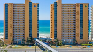 Un hôtel de grande hauteur doté de nombreux balcons, offrant une vue sur la plage et relié à un pont piétonnier.