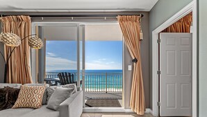 Un balcon avec vue sur l’océan, un canapé gris avec des coussins à motifs et une porte sur la droite.