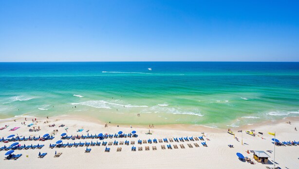 Une plage bordée de rangées de chaises longues et de parasols, un ciel bleu azur et l’océan.