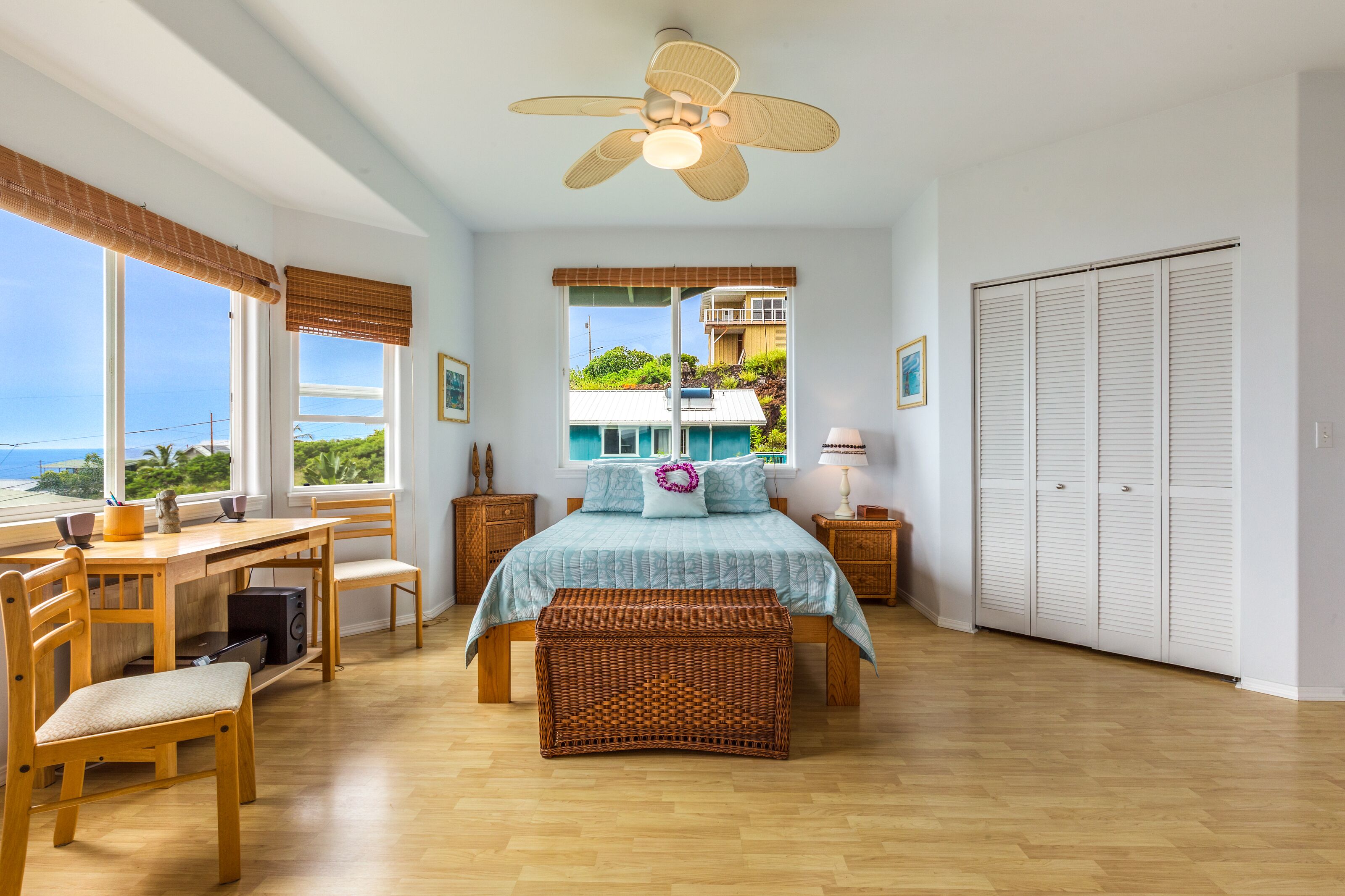 Une chambre avec un lit, un bureau en bois, des chaises, un banc en osier, un ventilateur de plafond et une vue sur une maison et l’océan.