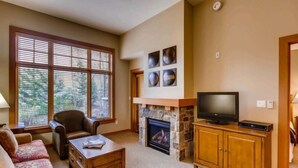 A living room with a fireplace, a television on a wooden cabinet, a sofa, and a window with blinds. - Capitol Peak Lodge 3123 (Snowmass Village)