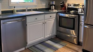 A modern kitchen with white cabinets, stainless steel appliances, a window with a floral valance, and a patterned rug. - Cozy and Convenient! (Cedar)
