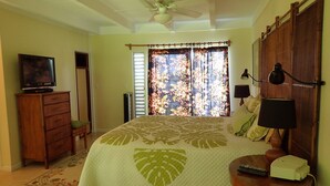 A bedroom with a bed, a dresser, a television, a ceiling fan, a window with curtains, and a nightstand with a lamp and remote control. - Beautiful 2/2 Townhouse in Princeville, Free WiFi, Cable TV, BBQ, beach gear (Princeville)