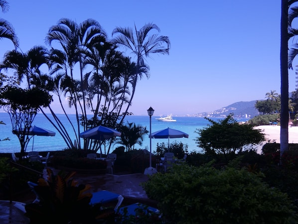 Une terrasse au bord de la plage, agrémentée de parasols, de palmiers et offrant une vue sur l’océan et un yacht.