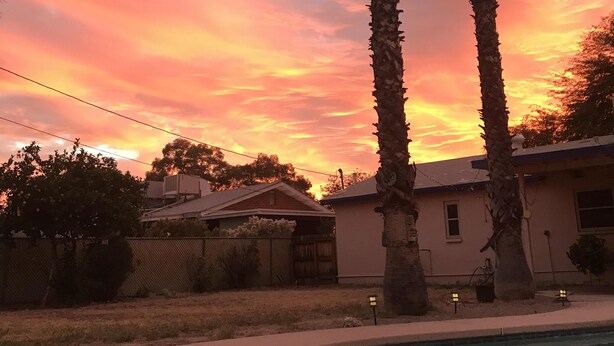 Une piscine avec un ciel au coucher du soleil, des palmiers et une maison en arrière-plan.
