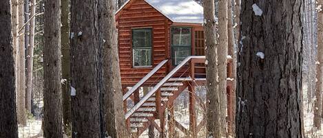 A wooden cabin with a staircase leading to the entrance, surrounded by trees and snow.