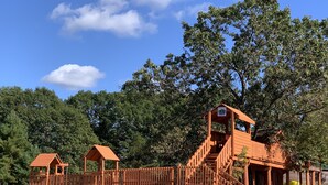 A wooden playground structure with slides and climbing areas, surrounded by trees and grass. - Vacation Rental in Westfield/Oxford, Wis., with acres of nature … (Oxford)
