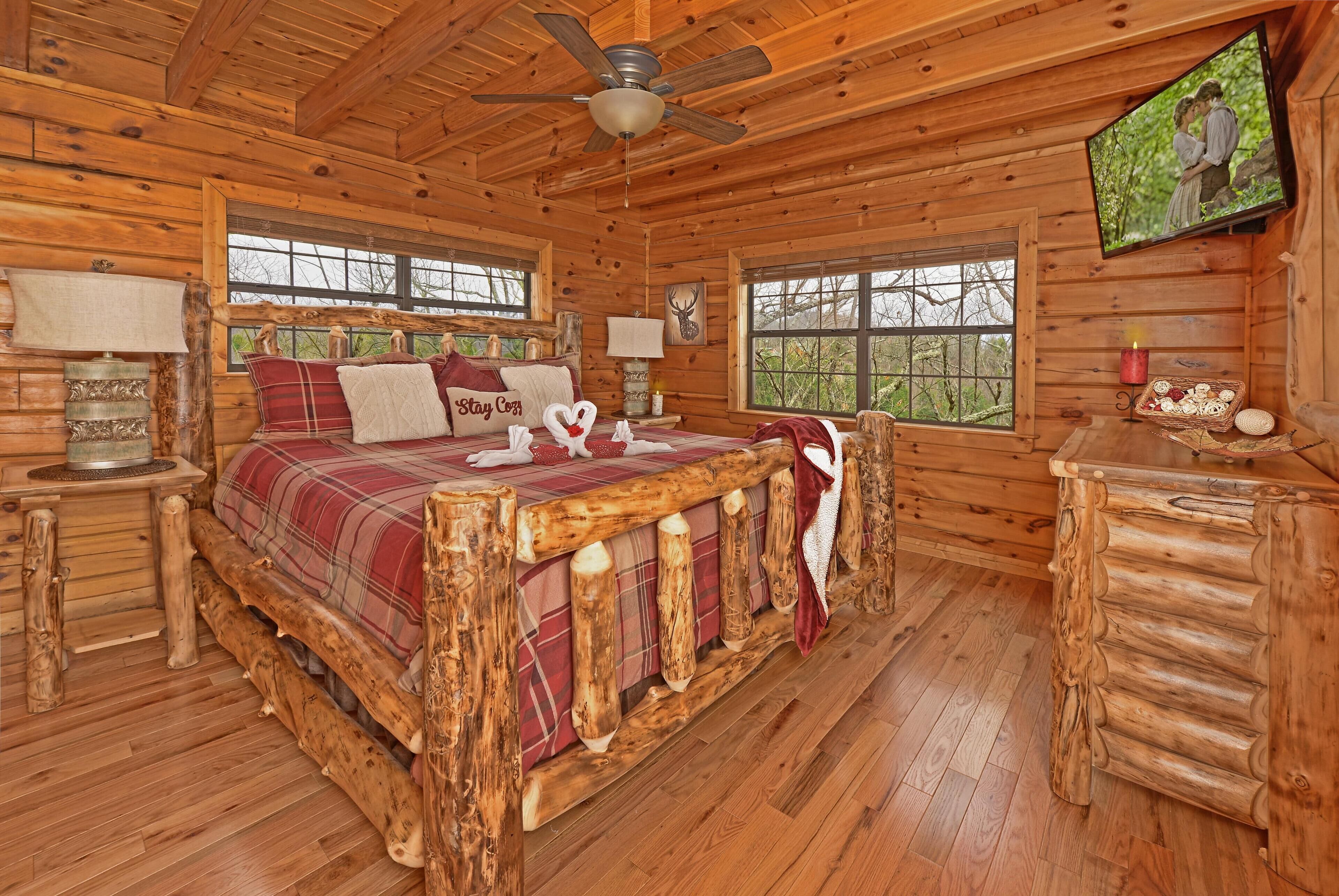 A log cabin bedroom with a wooden bed, a ceiling fan, a television, and a window with a view of trees.