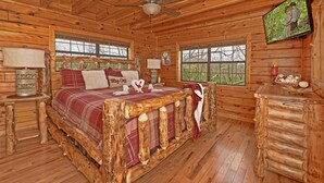 Ein Schlafzimmer in einer Blockhütte mit einem Holzbett, einem Deckenventilator, einem Fernseher und einem Fenster mit Blick auf Bäume.