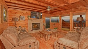 Ein Holzhäuschen im Inneren mit Kamin, Ledersofas, einem Fernseher und Blick auf die Berge.