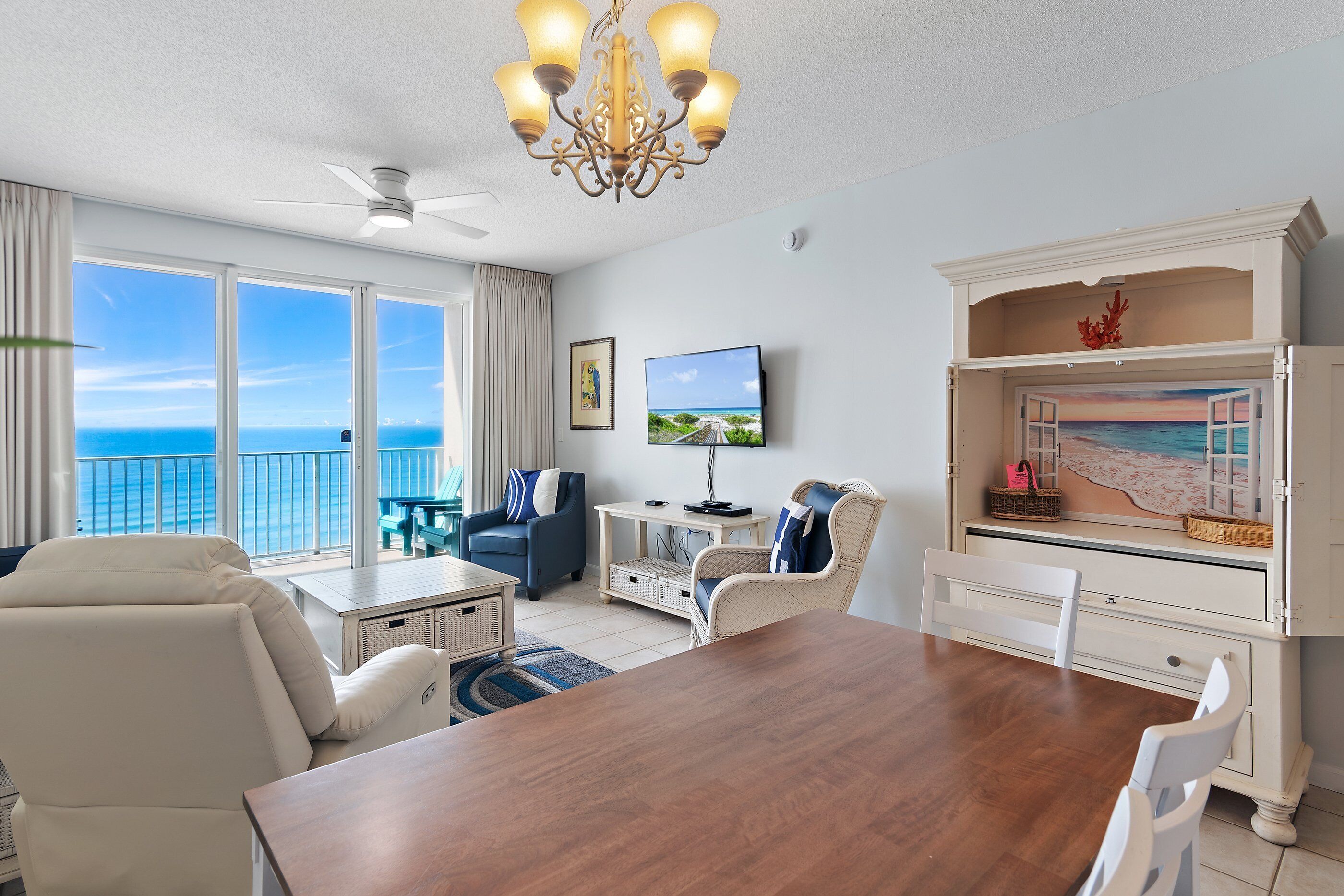 A spacious living room with a large wooden table, white chairs, a ceiling fan, and a television.