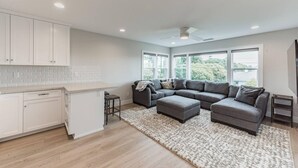 A modern kitchen with white cabinets and a marble countertop, a gray sectional sofa with a patterned rug, and large windows offering a view of greenery.