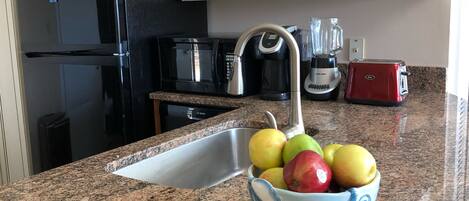 Una cocina con encimera de granito, un fregadero, un lavavajillas Bosch, un refrigerador negro, un tostador rojo y un bol de manzanas.