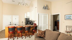 A compact living space with a kitchenette, a dining area with bar stools, a sofa with a patterned throw, and a view of the outdoors through a door. - Romantic SML Penthouse, Bernard’s Landing. Smith Mountain Lake 3 Pools (Moneta)