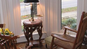 A room with a view of a street, a wooden rocking chair, a small table with a lamp, and a chair with a floral arrangement. - Historic Home - Victorian Stage Coach Inn (Russellville)