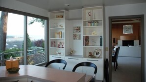A modern room with a large window, white shelving with decorative items, a dining area with a table and chairs, and a view of greenery outside. - 'Harbor View Grand'—Grand View, Home, Hot Tub, Sauna, and Concert Grand Piano. (Oak Harbor)