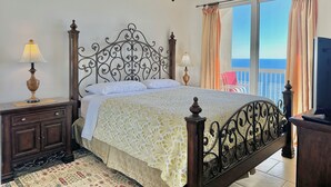 A bedroom with a bed, two bedside tables with lamps, a ceiling fan, a large window with curtains, and a view of the sea.
