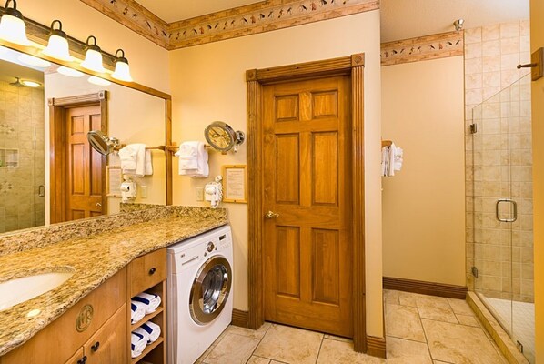 A hotel bathroom with a granite countertop, a washing machine, a wooden door, a shower, and a mirror with lights.
