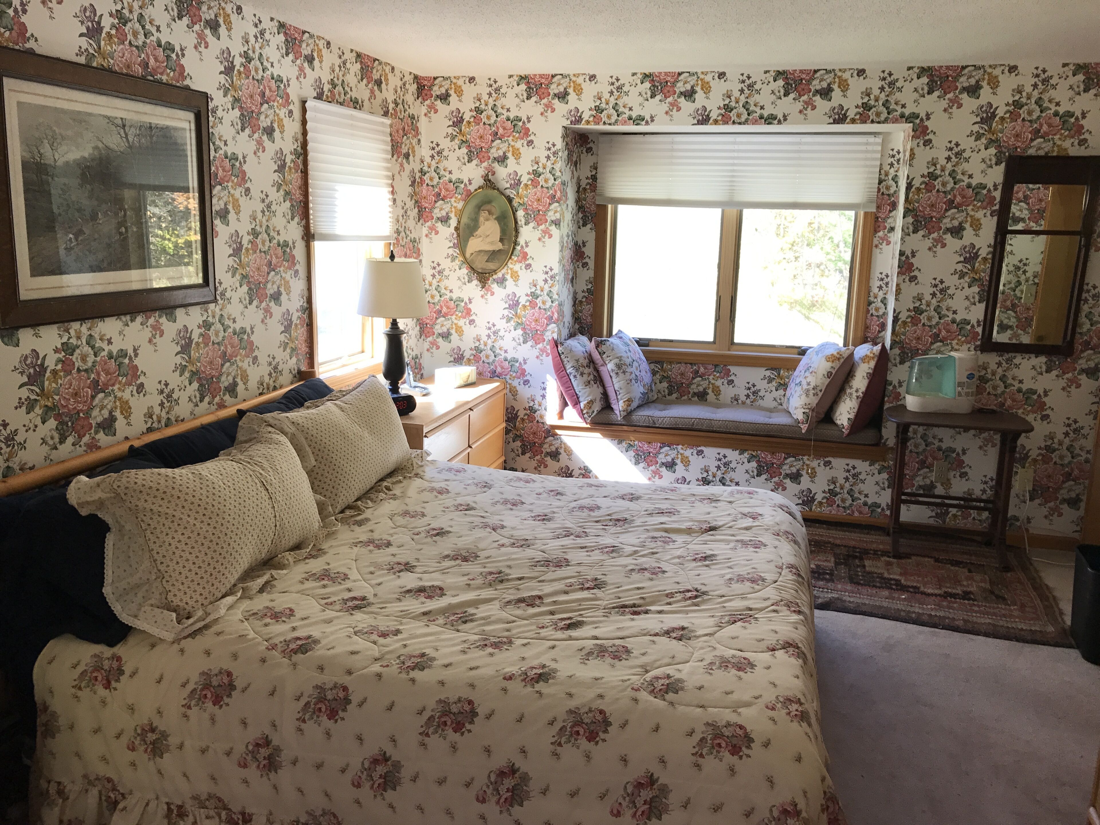 A bedroom with floral wallpaper, a bed with floral bedding, a window seat with cushions, a bedside table with a lamp, and a framed picture on the wall.