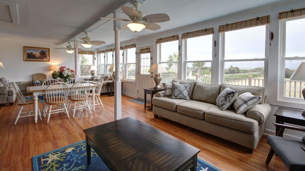 A spacious living room with a beige sofa, wooden dining table, and chairs. There are large windows with blinds, a ceiling fan, and a view of the outdoors. - Oceanfront Vintage, 1960s Cottage! Very Cozy w/Large Backyard, Ample Space, & Prime Location! (Isle of Palms)
