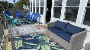 A covered deck with a large tropical leaf-patterned rug, blue Adirondack chairs, and a wicker sofa with blue cushions. - Oceanfront Vintage, 1960s Cottage! Very Cozy w/Large Backyard, Ample Space, & Prime Location! (Isle of Palms)