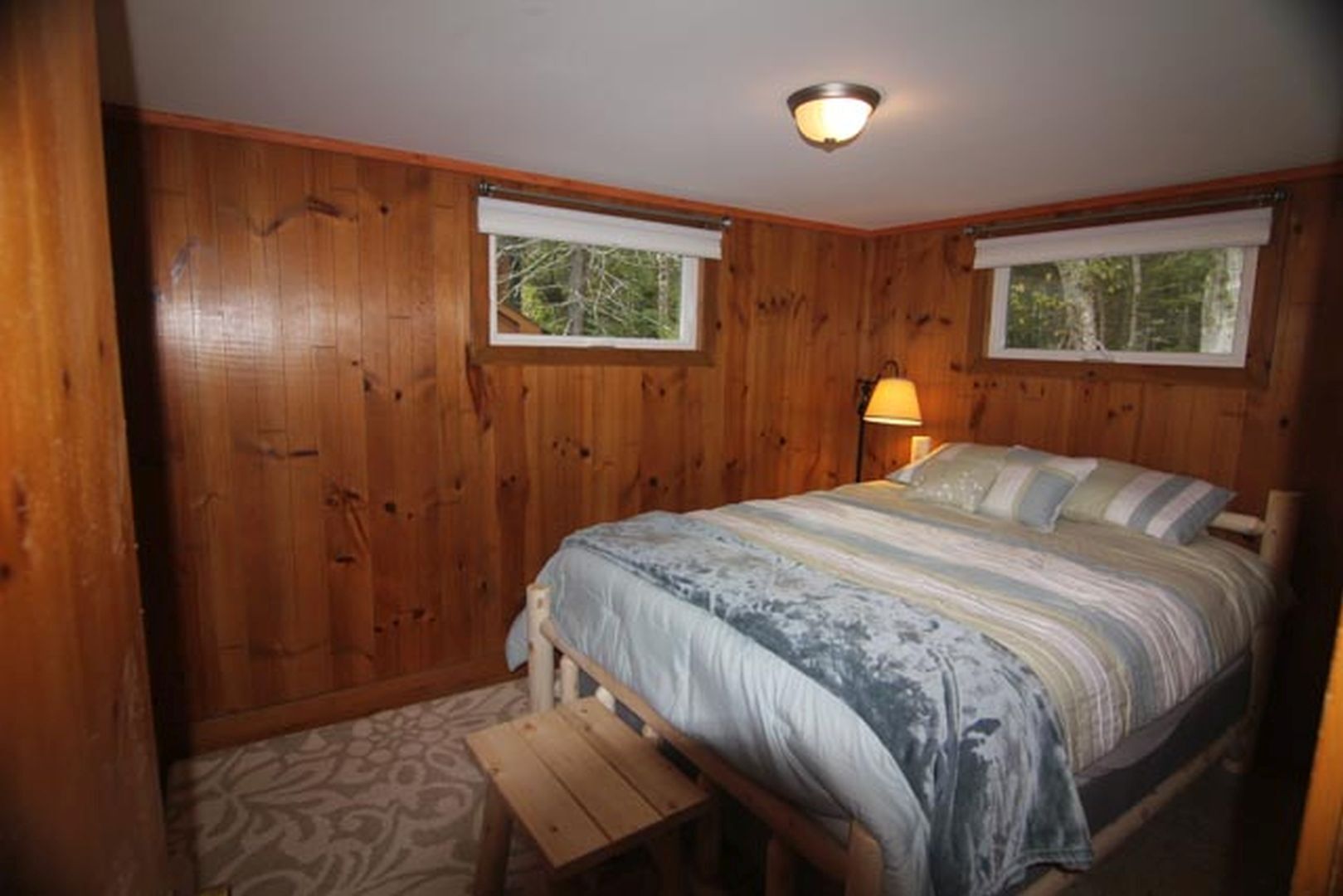 A wooden-paneled bedroom with a bed, two windows, a nightstand, and a lamp.
