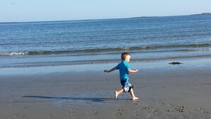 Un enfant court sur une plage de sable, laissant des empreintes derrière lui. Le ciel est dégagé et la mer est calme à l'arrière-plan.
