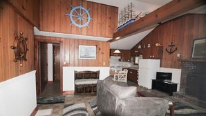 Un salon chaleureux avec un décor intérieur en bois, une grande roue de navire accrochée au mur, une cheminée et un coin repas avec une table et des chaises.