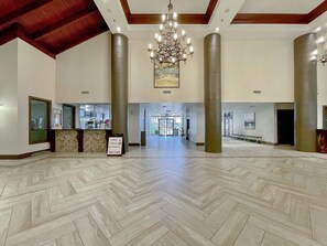 A spacious lobby with a herringbone floor pattern, a central chandelier, and a reception desk with a sign advertising coffee, donuts, and ice cream.