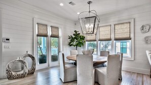 Un comedor con mesa de madera y seis sillas, ventanas grandes con persianas, una lámpara colgante y una planta en maceta.