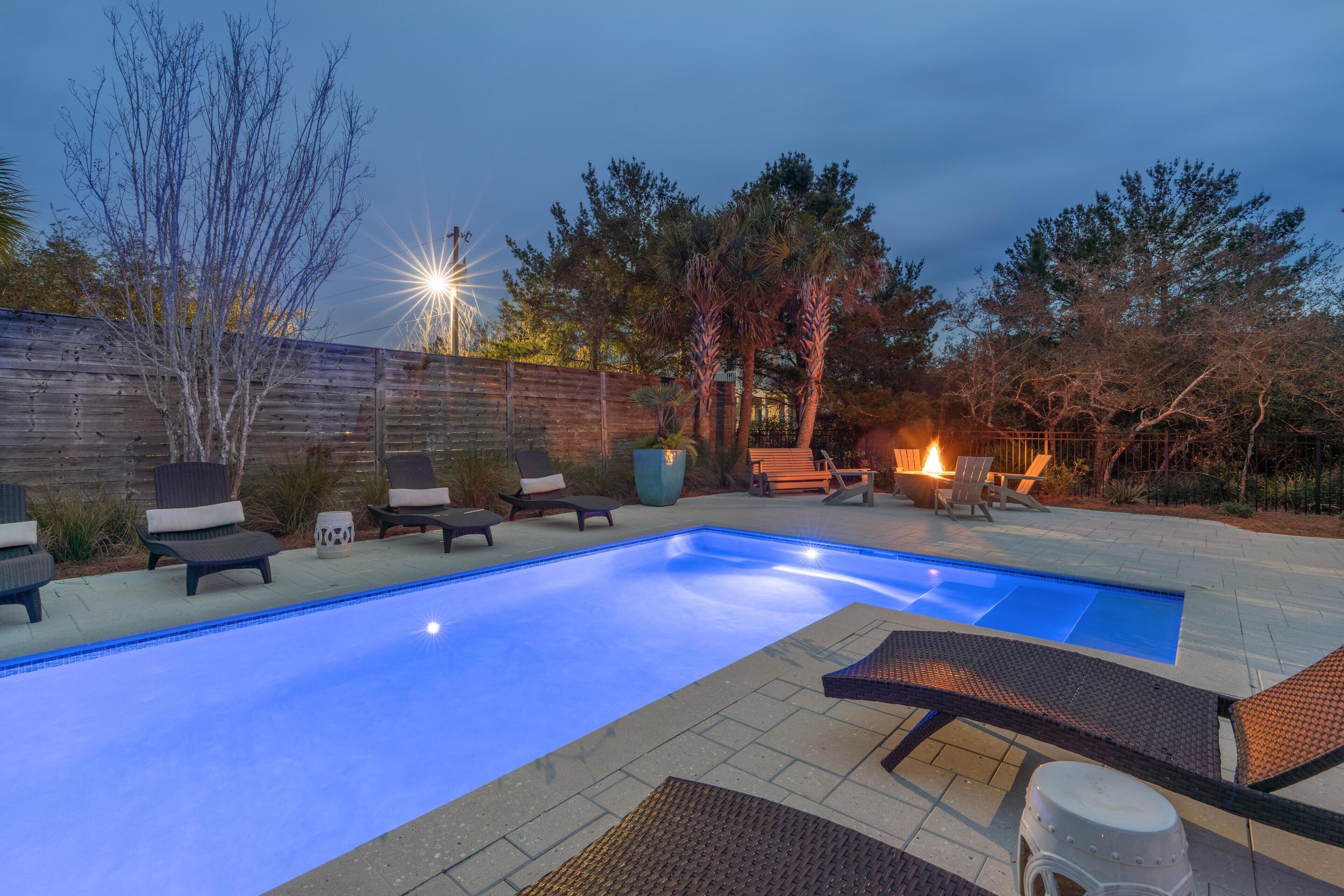 A swimming pool with lounge chairs and a fire pit area, surrounded by trees and a wooden fence.