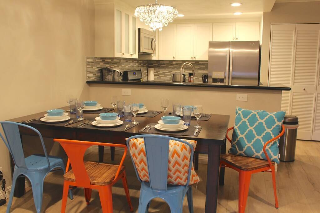 A dining area with a black table set for four, blue and orange chairs, a chandelier, and a kitchen in the background.
