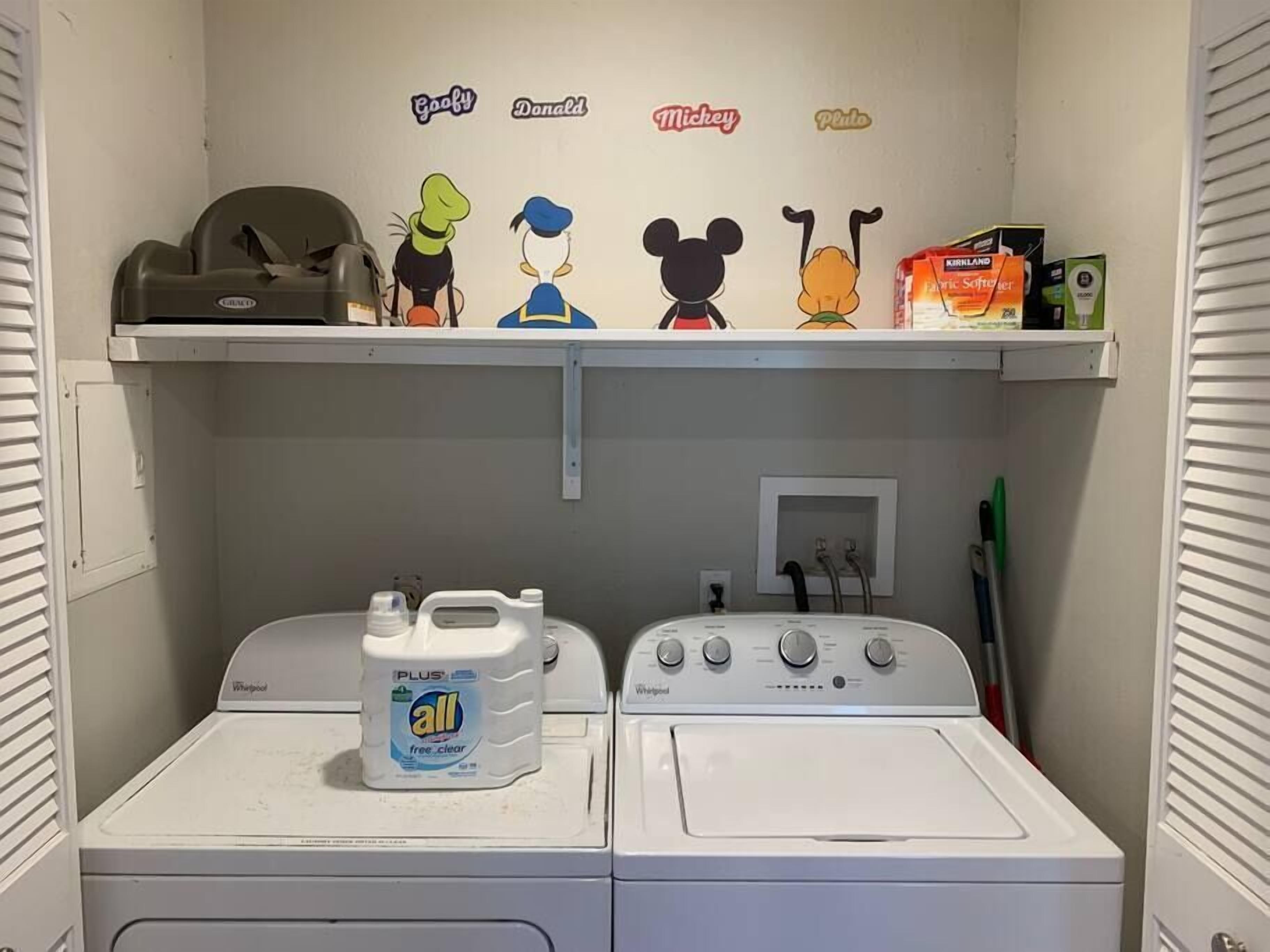 A laundry room with two Whirlpool washers, a shelf with cartoon character decorations, and a bottle of Plus all free clear detergent.