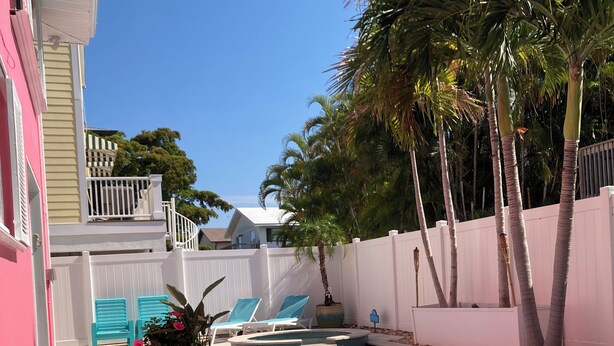 Una piscina con agua cristalina, rodeada de una cerca blanca, palmeras y un edificio rosa.