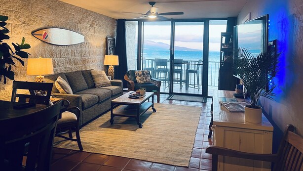 A living room with a stone wall, a ceiling fan, a large flat-screen TV, and a view of the sea through a sliding glass door. -  Crew Rest Maui - 2BR/2BA, Oceanfront, Superior Views, A/C, sleeps 6 (Wailuku)