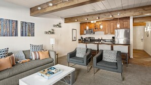 Una sala de estar moderna con un sofá gris, dos sillones y una mesa de centro. Se ve una cocina en el fondo con gabinetes de madera y electrodomésticos de acero inoxidable.