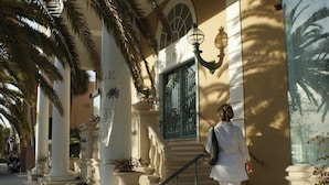 Uma pessoa caminhando em direção a um edifício com uma grande janela e uma palmeira em primeiro plano.
