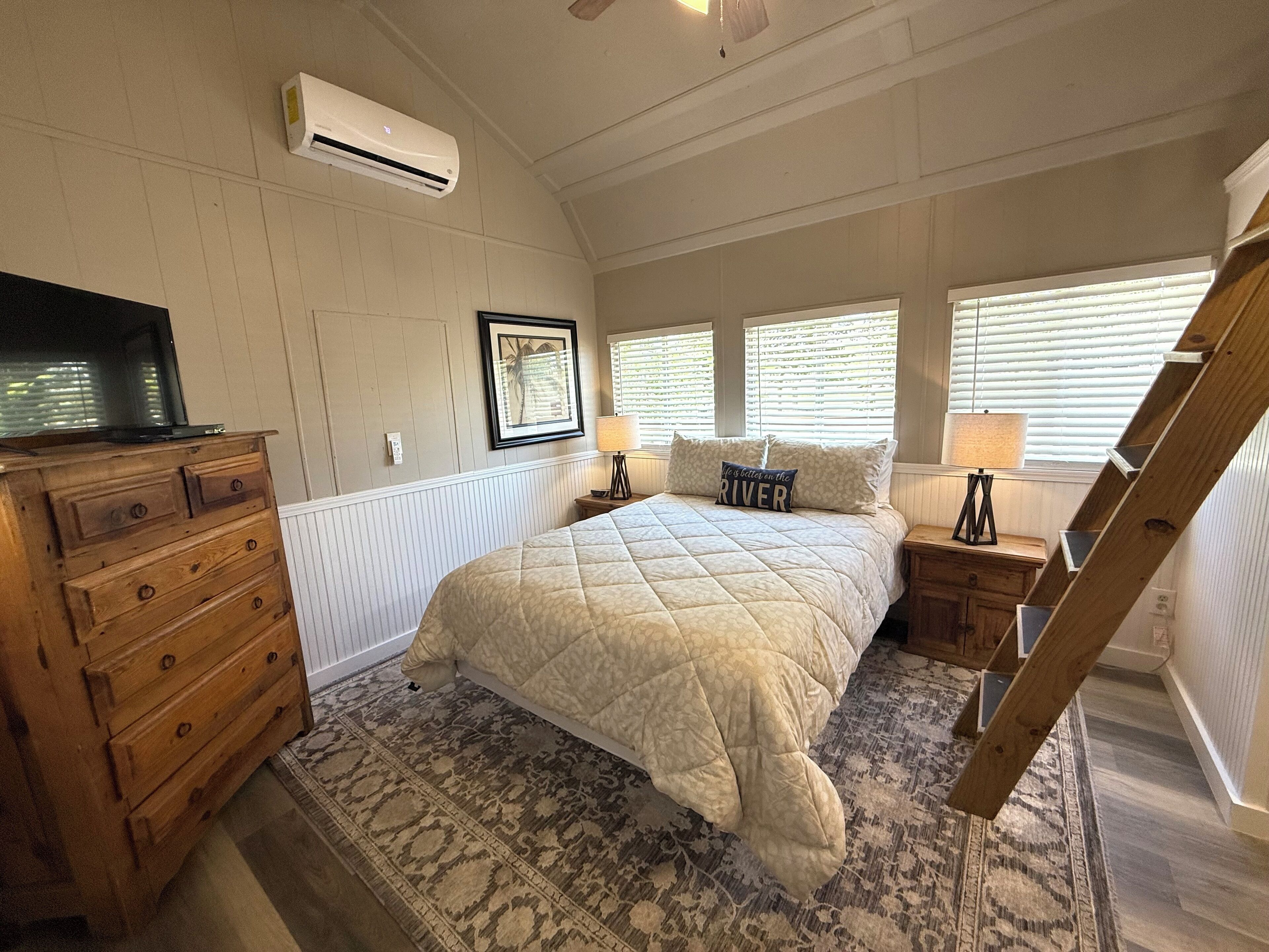 A bedroom with a bed, dresser, air conditioning unit, framed picture, bedside lamps, and a window with blinds.