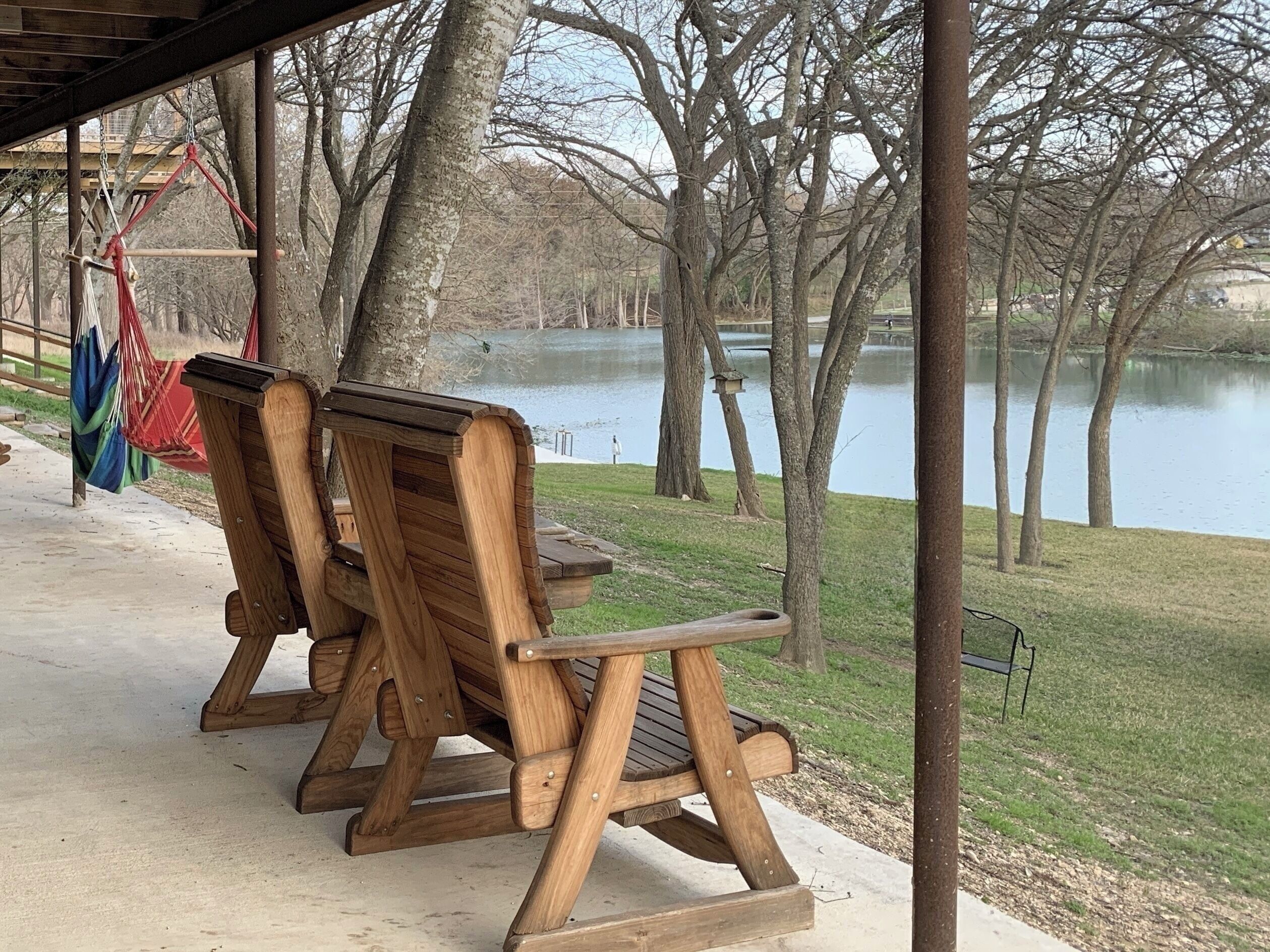 A wooden bench with a backrest and armrests, a hammock, and a view of a lake surrounded by trees.