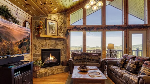A cozy living room with a stone fireplace, wooden furniture, and large windows offering a view of the outdoors. - 4 bedroom 4 bathroom sleeps 14 outdoor pool access great mountain views and free internet access (Sevierville)