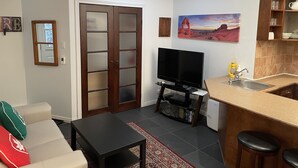 A living room with a TV, sofa, coffee table, and a kitchen area with a sink and bar stools. - Cute and  Cozy basement unit in Tremblant (Mont-Tremblant)