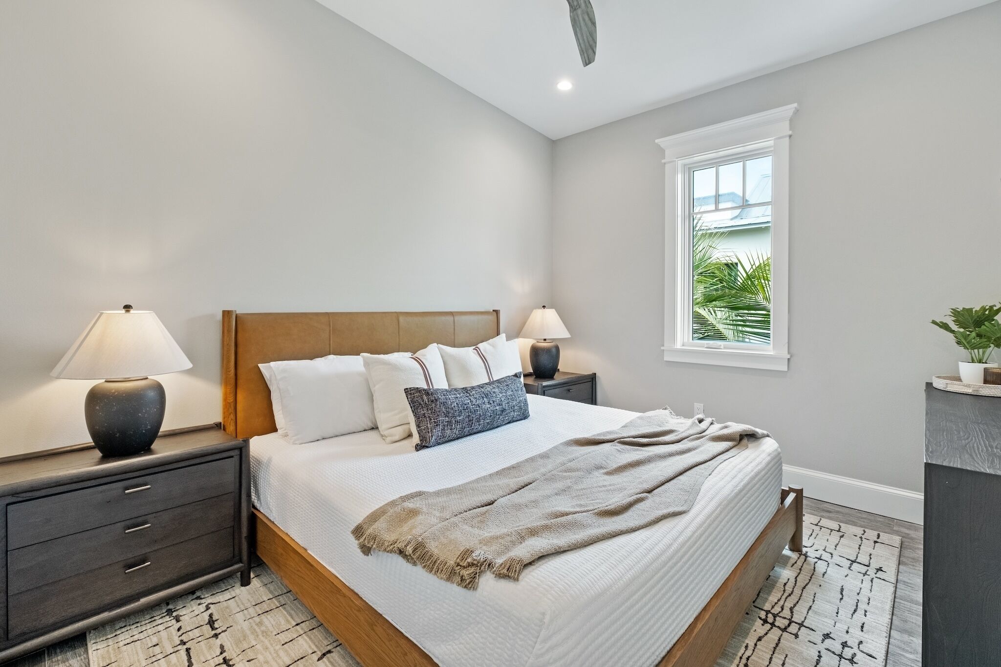 A modern bedroom with a wooden bed, two bedside tables with lamps, a large window with a view, and a small plant on a side table.