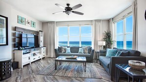 Un salón amplio con un gran ventanal con vistas al océano, un ventilador de techo y un televisor de pantalla plana sobre un mueble blanco.