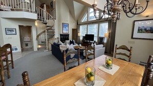 A spacious living area with a wooden dining table, two chairs, a sofa, and a television. A staircase leads to an upper level.